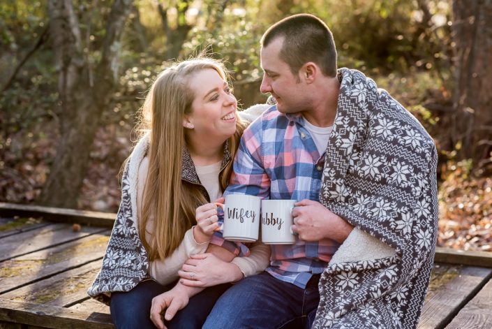 Couple snuggles under blanket on achilly morning with coffee mugs for engagement photos in Crump Park, Glen Allen, VA