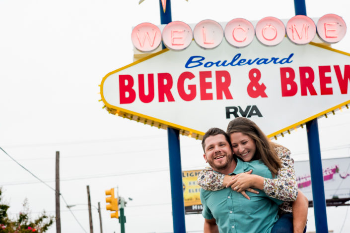 Engagement session couple gives piggy back ride in front of Boulevard Burger and Brew sign in downtown Richmond, VA